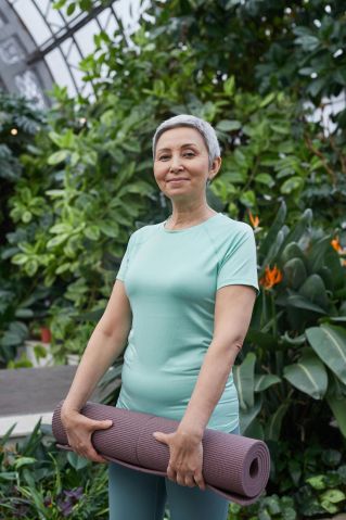 Smiling senior woman with yoga mat in a vibrant greenhouse setting, promoting well-being.